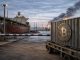 Bitcoin container halted at a port checkpoint as smoke rises in the distance, reflecting how escalating Iran war fears are weighing on the crypto market